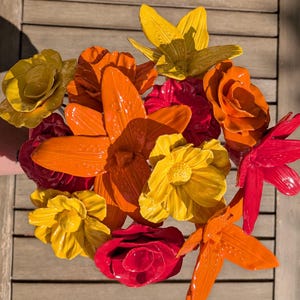 May include: A vibrant bouquet of painted metal flowers in shades of yellow, orange, and red. The arrangement includes various flower types, such as lilies and roses, with glossy finishes. The flowers are arranged on a wooden surface.