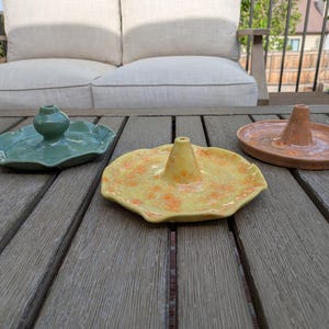 May include: Three ceramic incense holders in shades of green, yellow, and orange. Each holder has a unique design, with a central cone or vase shape. The holders are placed on a wooden table, showcasing their handmade quality.