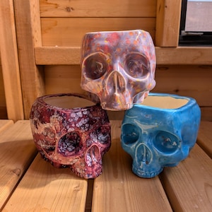 Pode incluir: Três vasos de cerâmica em forma de caveira em tons de vermelho, azul e laranja. Os vasos têm um acabamento brilhante e estão dispostos sobre uma superfície de madeira. Os vasos de caveira são um item decorativo único.