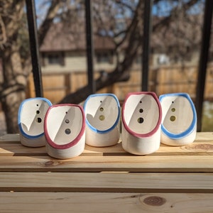 Puede incluir: Cinco pequeñas macetas de cerámica blancas con detalles azules y rosas. Las macetas están dispuestas en fila sobre una superficie de madera. Cada maceta tiene un pequeño agujero en el fondo para el drenaje.