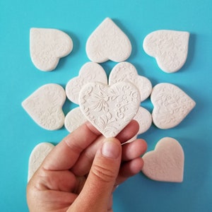 Op de afbeelding: Een hand houdt een wit hartvormig keramiek ornament vast met een bloemenpatroon. Het ornament is omringd door andere witte hartvormige ornamenten op een turquoise achtergrond.
