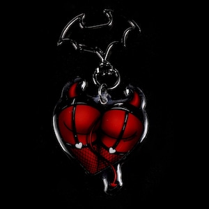 May include: A heart-shaped keychain featuring a devilish design. The keychain has a red heart with black accents, fishnet stockings, and a small devil tail. The top of the keychain has a bat-shaped design and a silver chain.