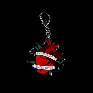 May include: A keychain featuring a red anatomical heart design with a banner that reads "A heart's a heavy burden." The heart is surrounded by green leaves and is attached to a silver keyring.