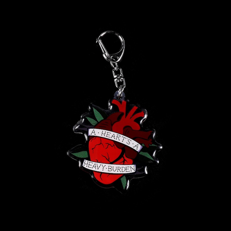 May include: A keychain featuring a red anatomical heart design with a banner that reads "A heart's a heavy burden." The heart is surrounded by green leaves and is attached to a silver keyring.