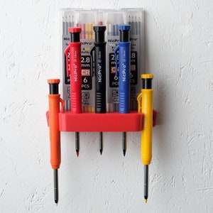 May include: A red wall-mounted holder with four mechanical pencils. The pencils are orange, red, black, and yellow. Each pencil has a clear plastic barrel with a black tip and a metal clip. The pencils are labeled "NicPro" and "2.8 mm CR 6 PCS".