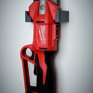 May include: A red and black Milwaukee M12 tool is mounted on a gray holder against a white wall. The tool has a black handle and a red body with the Milwaukee logo. A black cord extends from the bottom.