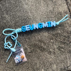 Könnte beinhalten: Ein hellblauer Schlüsselanhänger mit dem Namen SEUNGMIN, der in abwechselnd hell- und dunkelblauen Blockbuchstaben auf einer Schnur steht. Daran befestigt ist ein kleiner durchsichtiger Kunststoffbeutel mit einem Foto, einer Blumenperle und einem Bären.