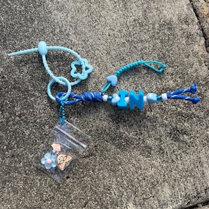 Könnte beinhalten: Ein blauer Schlüsselanhänger mit einer geflochtenen Kordel, den Buchstaben IN, einem hellblauen Silikonstern und einem kleinen durchsichtigen Plastikbeutel mit einer blauen Blumenperle und einem beigen Tieranhänger an einer Kugelkette.