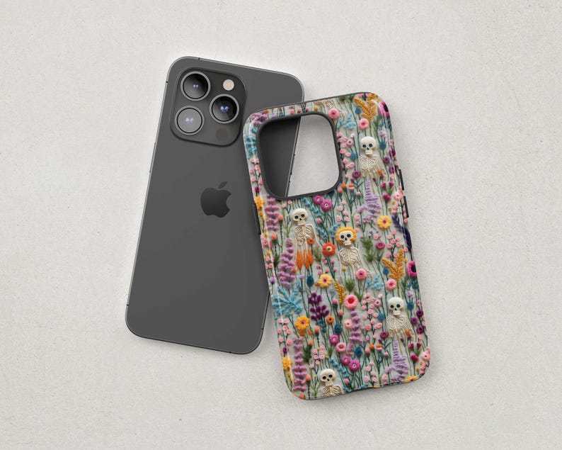 May include: A dark gray smartphone next to a phone case with a floral and skeleton design. The case features a colorful pattern of flowers and small skeletons.