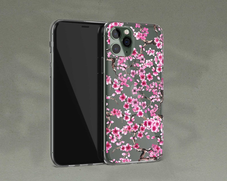 Puede incluir: Una funda transparente para tel&eacute;fono con un dise&ntilde;o floral que presenta flores de cerezo rosas sobre un fondo gris. La funda est&aacute; dise&ntilde;ada para un iPhone con una c&aacute;mara de triple lente.