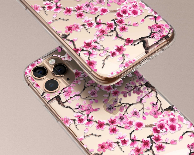 Puede incluir: Una funda transparente para tel&eacute;fono con un dise&ntilde;o floral con flores de cerezo rosas sobre un fondo dorado. La funda est&aacute; dise&ntilde;ada para un iPhone con una c&aacute;mara de triple lente.