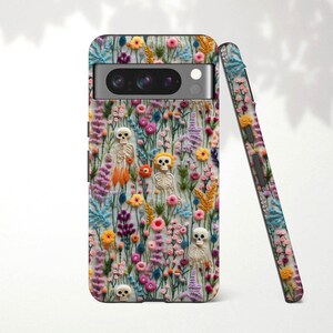 May include: A phone case featuring a colorful floral design with embroidered skeletons. The case is covered in a variety of flowers in shades of pink, yellow, and blue. A second case is leaning against the first, showing the same design.