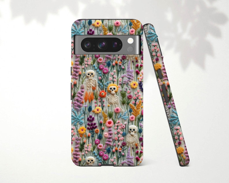 May include: A phone case featuring a colorful floral design with embroidered skeletons. The case is covered in a variety of flowers in shades of pink, yellow, and blue. A second case is leaning against the first, showing the same design.