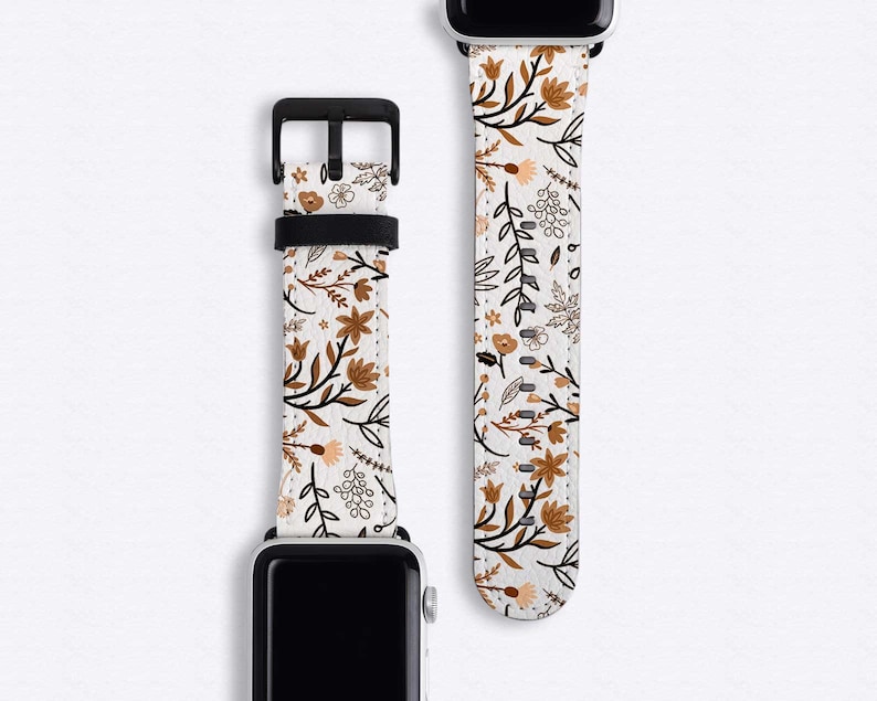 May include: A white Apple Watch band with a floral pattern featuring brown and black leaves and flowers. The band is made of leather and has a silver buckle.