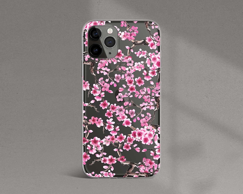 Puede incluir: Una funda transparente para tel&eacute;fono con un dise&ntilde;o de flores de cerezo rosas. La funda tiene un fondo negro y presenta un patr&oacute;n de flores rosas y ramas.