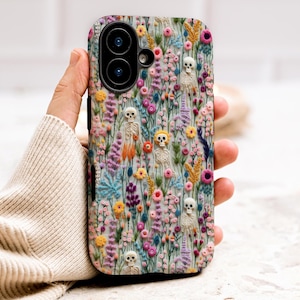 May include: A phone case with a floral design and embroidered skeletons. The case has a light blue background with pink, yellow, and purple flowers. The skeletons are white and orange, adding a unique touch to the design.