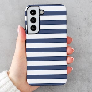 Peut inclure: Une coque de smartphone à rayures blanches et bleu marine. La coque présente un module caméra avec trois objectifs et un flash. La coque est tenue dans une main.