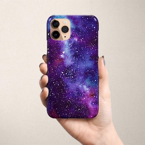Puede incluir: Una funda para teléfono con un diseño de galaxia. La funda es de color morado, azul y negro con estrellas blancas. La funda del teléfono tiene un recorte para el objetivo de la cámara.