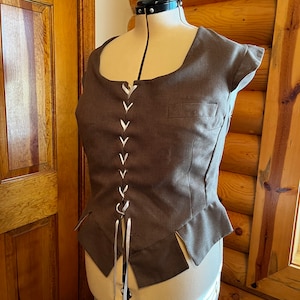 May include: A brown linen bodice with a white lace-up front and a fitted silhouette. The bodice has a square neckline and short sleeves.