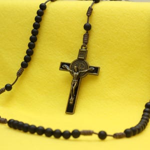 May include: A rosary with black beads and a bronze crucifix. The crucifix features a detailed depiction of Jesus Christ. The beads are strung on a brown cord, and the background is a solid yellow.