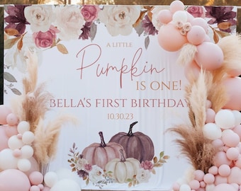 Op de afbeelding: Een witte achtergrond met een bloemenontwerp en drie pompoenen. De tekst "A Little Pumpkin Is One! Bella's First Birthday 10.30.23" is op de achtergrond gedrukt. Op de voorgrond zijn roze en witte ballonnen te zien.