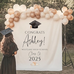 May include: White graduation backdrop with black text that reads "Congrats Ashley! Westlake High School Class of 2025". A black graduation cap with a gold tassel is above the text. A gold and white balloon arch is above the backdrop.