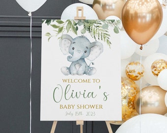 May include: A baby shower welcome sign featuring a watercolor illustration of a gray elephant with green foliage. The sign reads "Welcome to Olivia's Baby Shower July 15th, 2023" in green and gold lettering. The sign is displayed on a wooden easel.