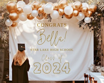 May include: A white backdrop with gold lettering that reads "Congrats Bella! Star Lake High School Class of 2024". A person wearing a black graduation gown and cap stands in front of the backdrop. A garland of gold and white balloons hangs above the backdrop.