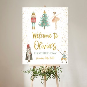May include: A welcome sign for Olivia's first birthday, featuring watercolor illustrations of a nutcracker, Christmas tree, ballerina, and other characters. The sign has gold text and accents, with the date December 25th, 2022.