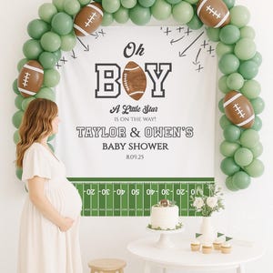 May include: A baby shower scene with a white backdrop, green balloons, and football decorations. The banner reads "Oh Boy" and "Taylor & Owen's Baby Shower." A pregnant person stands in front of the banner. A cake and cupcakes are on a white table.