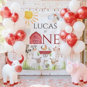 May include: A white banner with red and black accents reads "Lucas is One". The banner features a red barn, farm animals, and a red wagon. The banner is surrounded by red and white balloons.