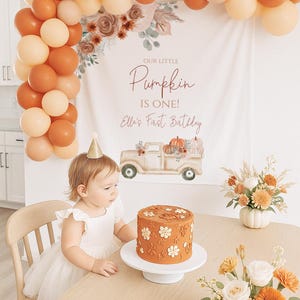 May include: A first birthday celebration with a pumpkin theme. A young child in a white dress and gold party hat sits at a table with a cake, floral arrangements, and a balloon arch in shades of orange and cream. A banner reads "Our Little Pumpkin is One!"