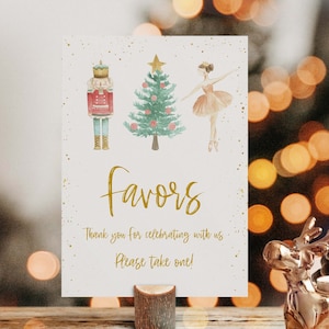 Pode incluir: Um cartão branco com letras douradas que dizem "Favors" e "Obrigado por celebrar conosco. Por favor, pegue um!" O cartão é decorado com ilustrações de aquarela de um quebra-nozes, uma árvore de Natal e uma bailarina. O cartão está em um suporte de madeira.