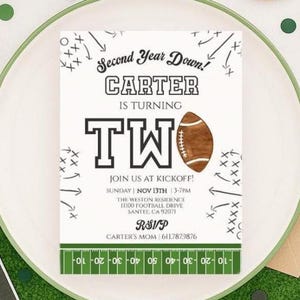 Puede incluir: Una tarjeta de invitación blanca con temática de fútbol. La tarjeta dice "Second Year Down! CARTER IS TURNING TWO" con un gráfico de fútbol. Incluye detalles del evento: fecha, hora, lugar e información de RSVP. La tarjeta está en un plato blanco.