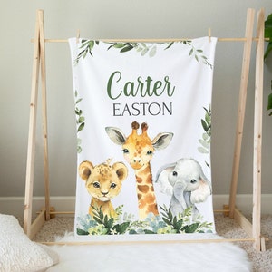 May include: A white baby blanket with a green leafy border. The blanket features a giraffe, lion, and elephant with the names "Carter Easton" printed on the blanket.