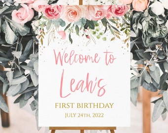 May include: A white welcome sign with pink floral accents and gold glitter. The sign reads "Welcome to Leah's First Birthday July 24th, 2022".