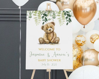 May include: A white sign on a wooden easel featuring a teddy bear illustration and foliage. The sign displays "Welcome to Jasmine & Aaron's Baby Shower July 15, 2023" in green and gold. White and gold balloons decorate the background.