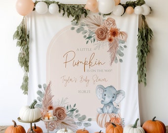 May include: A white backdrop with a peach-colored archway featuring a watercolor illustration of flowers, greenery, and a baby elephant sitting on a pumpkin. The text "A Little Pumpkin is on the way!" and "Taylor's Baby Shower 10.28.23" is printed on the backdrop. The backdrop is decorated with greenery and balloons.