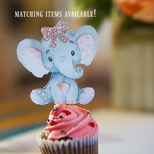 May include: A pink cupcake with a blue and white elephant topper. The elephant has a pink bow with glitter. The cupcake is decorated with red and white sprinkles. The text "MATCHING ITEMS AVAILABLE!" is visible in the background.