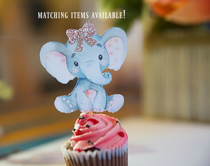 May include: A pink cupcake with a blue and white elephant topper. The elephant has a pink bow with glitter. The cupcake is decorated with red and white sprinkles. The text "MATCHING ITEMS AVAILABLE!" is visible in the background.