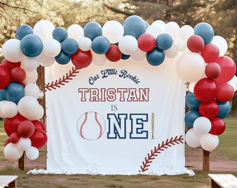 May include: A white banner with red and blue baseball themed decorations. The banner says "Our Little Rookie Tristan is One!" with a baseball and bat graphic. The banner is decorated with a garland of red, white, and blue balloons.