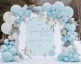 May include: A blue and white baby shower sign with the text "A Little Snowflake is on the way!" and "Rachel's Baby Shower 12.25.23". The sign is decorated with snowflakes and floral designs. The sign is surrounded by a blue and white balloon arch.