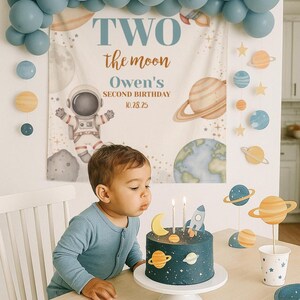 May include: A child blows out candles on a dark blue cake decorated with planets and a rocket. The cake sits on a white pedestal, with a banner that says "Two the moon" and a balloon arch in the background. The theme is a space-themed birthday party.
