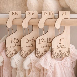 May include: Wooden closet dividers with age labels: NB (newborn), 3-6 months, 12-18 months, and 3T (toddler). Each divider has a floral design at the bottom. Pink baby clothes hang below. A light-colored basket is in the background.