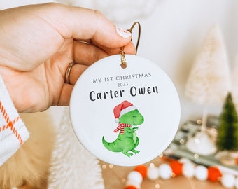 Könnte beinhalten: Runder weißer Weihnachtsbaumschmuck mit goldener Aufhängeschlaufe. Der Schmuck trägt den Text "MY 1ST CHRISTMAS 2023" und "Carter Owen". Ein grüner Dinosaurier mit Weihnachtsmütze und Schal ist ebenfalls abgebildet.