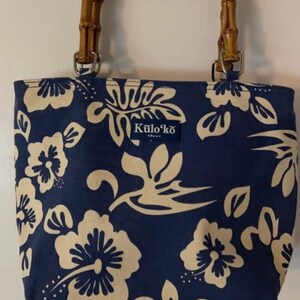 May include: A navy blue tote bag with a cream floral pattern and bamboo handles. The bag features the text "Kulo'ko" and is suspended from a gold-colored hook. The bag's design evokes a tropical theme.
