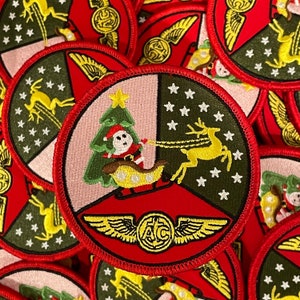 May include: A red, white, and green embroidered patch with a Christmas tree, reindeer, and a Santa Claus figure. The patch has a gold winged circle with the letters "ATC" in the center.
