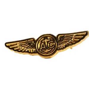 May include: Gold-colored metal pin with a pair of wings and a circular emblem in the center. The emblem has the letters "A.I.C." inscribed inside.