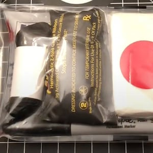 Könnte beinhalten: Eine transparente Plastiktüte mit einem Erste-Hilfe-Set. Das Set enthält ein hämostatisches Z-Falz-Gazeband, einen Verband, einen Marker und einen roten Kreis auf weißem Hintergrund.