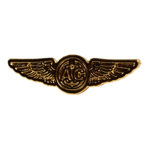 May include: A gold-colored metal pin with a pair of wings and a circle in the center. The circle has the letters "AC" inside.